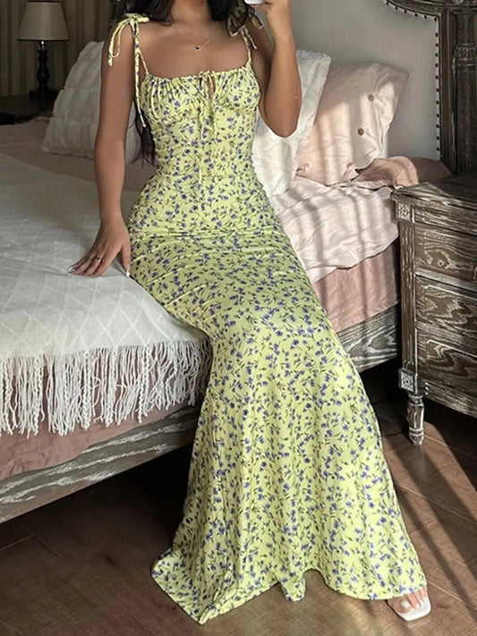 Woman in yellow floral maxi dress with tie straps, sitting on bed in cozy bedroom