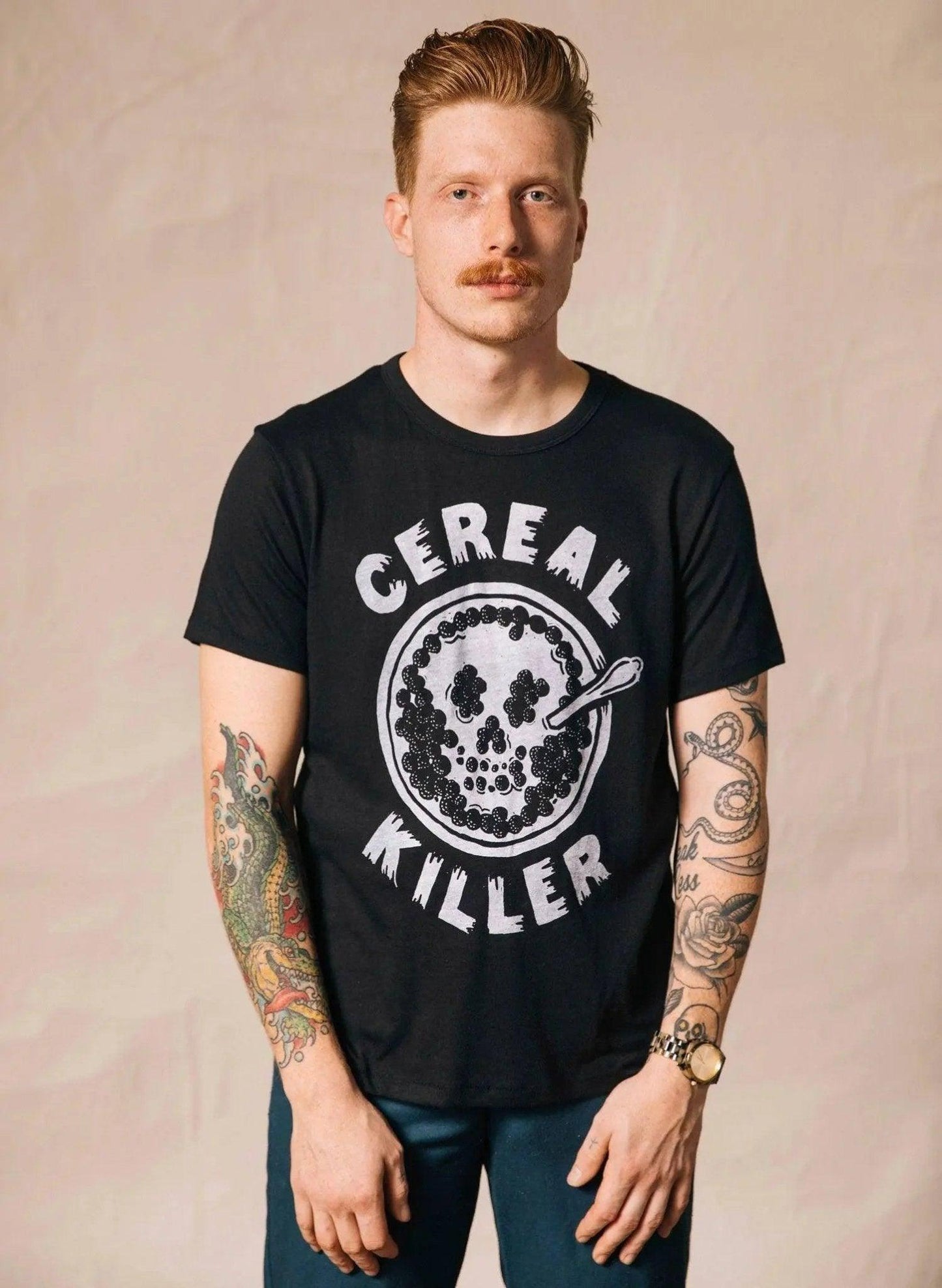 Man wearing black 'Cereal Killer' graphic tee with tattooed arms, casual fashion style