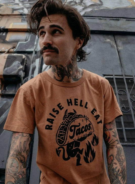 Man wearing 'Raise Hell Eat Tacos' graphic tee, tattooed arms, urban street style fashion