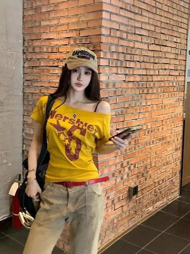Woman in yellow graphic tee, cap, and cargo pants leaning against brick wall — trendy streetwear fashion