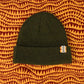 Olive green ribbed knit beanie with EKZO tag, styled on patterned fabric background