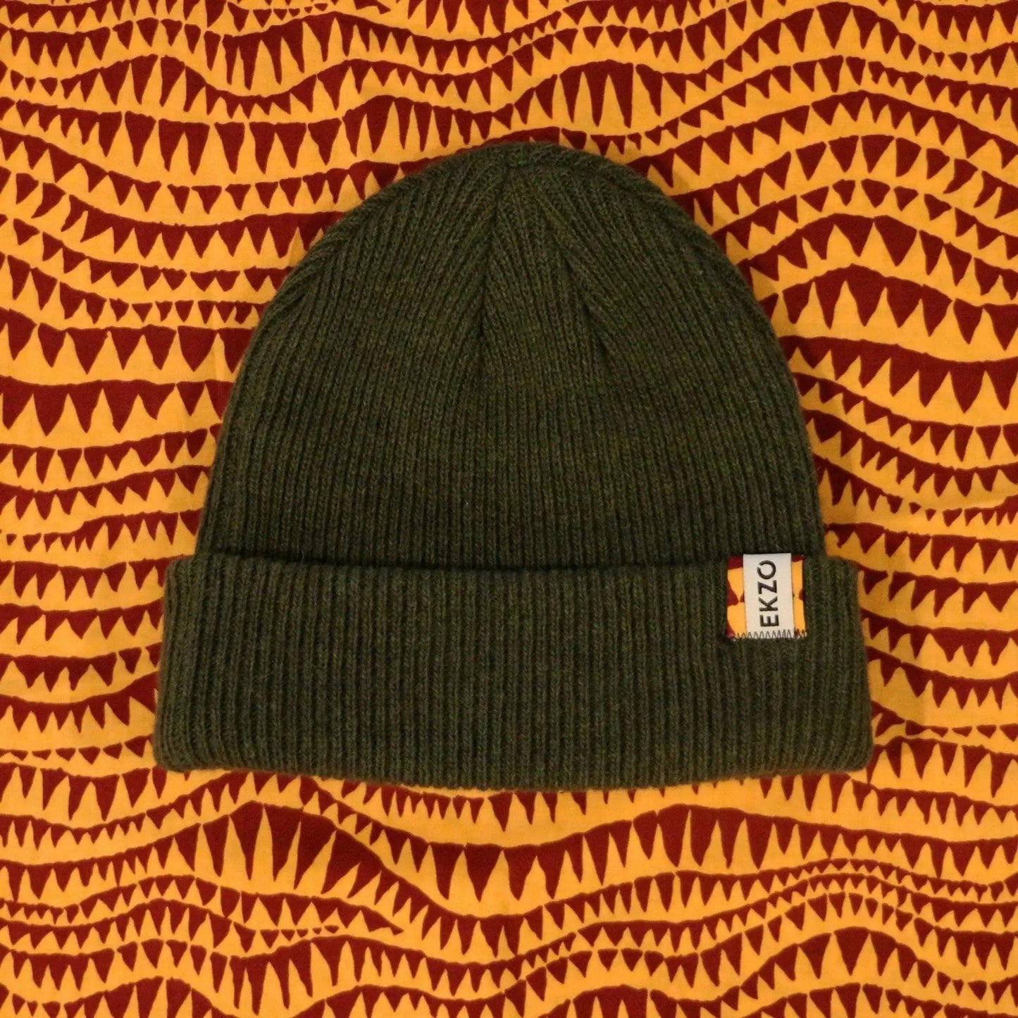 Olive green ribbed knit beanie with EKZO tag, styled on patterned fabric background