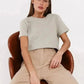 Woman in light gray crop top and beige pants, seated on brown chair, modern fashion look