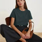 Woman in dark green t-shirt, black pants, and brown handbag, seated on wooden chair — stylish fashion outfit