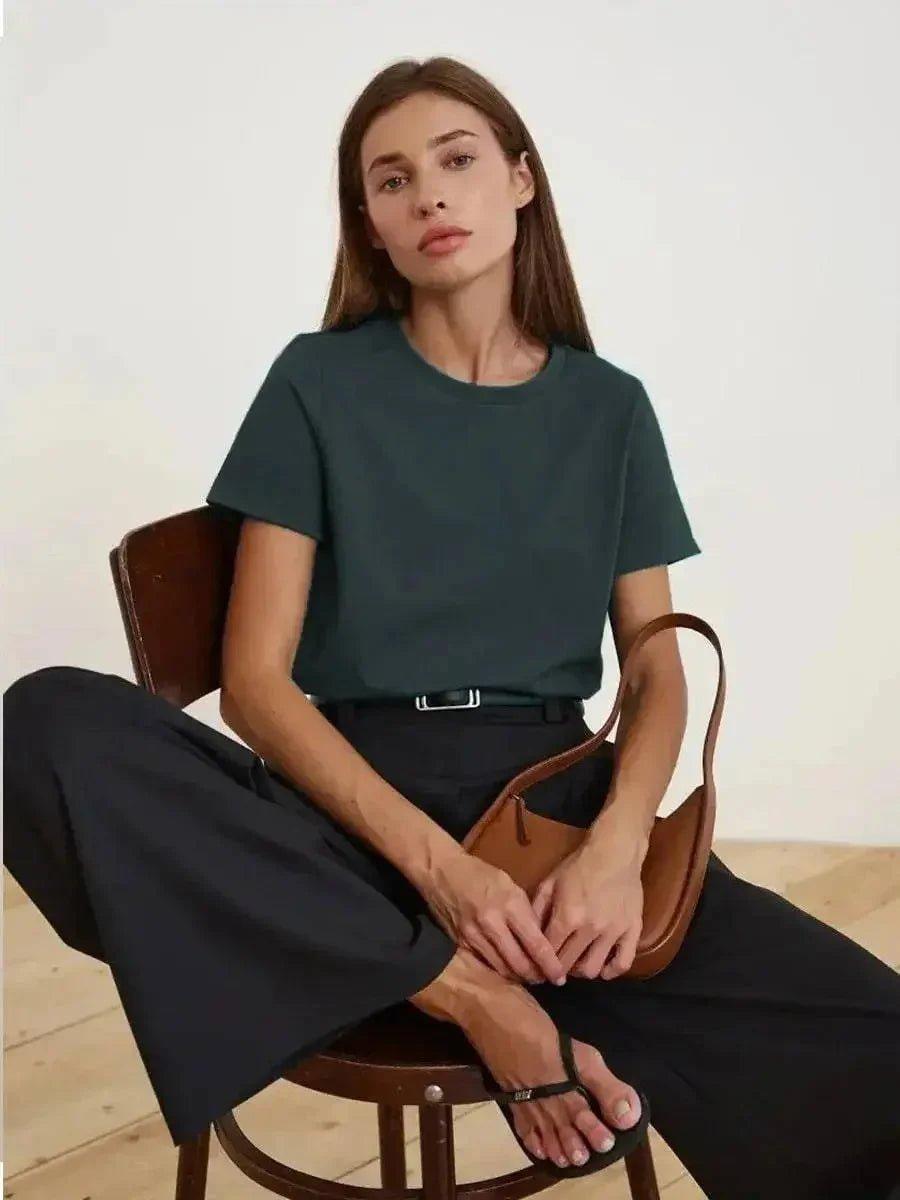 Woman in dark green t-shirt, black pants, and brown handbag, seated on wooden chair — stylish fashion outfit