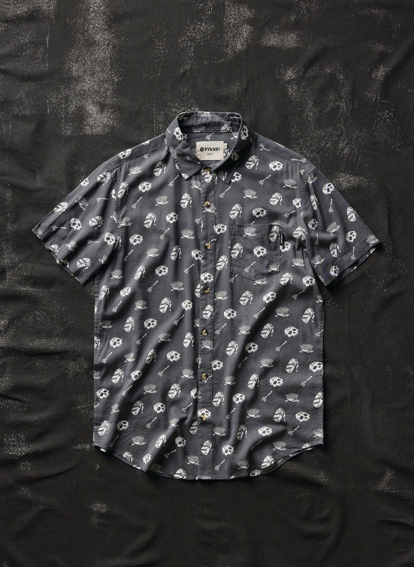Dark gray short sleeve button-up shirt with skull and bowl print, laid flat on textured black background