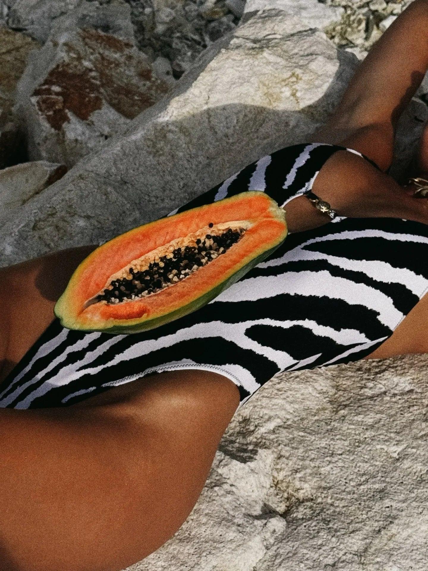 Woman in zebra print swimsuit holding fresh papaya on rocky beach, summer fashion vibe