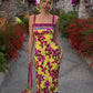 Woman in vibrant yellow floral midi dress with pink straps, holding matching bag, walking through garden path