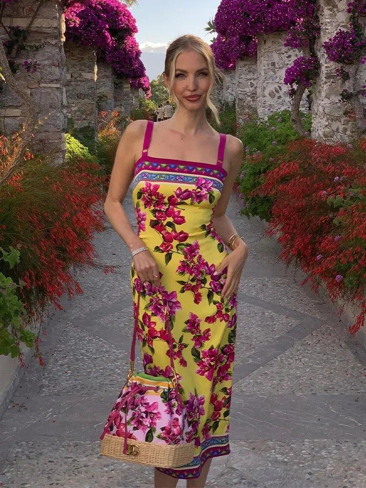 Woman in vibrant yellow floral midi dress with matching bag, standing on stone path surrounded by purple blooms