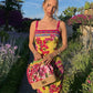 Woman in vibrant floral yellow dress with pink straps, holding woven bag, standing in garden