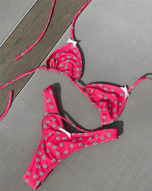 Pink floral bikini set with white daisy print, triangle top and thong bottom, laid on gray fabric