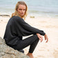 Woman in black long sleeve top and pants sitting on beach rock, coastal fashion outfit