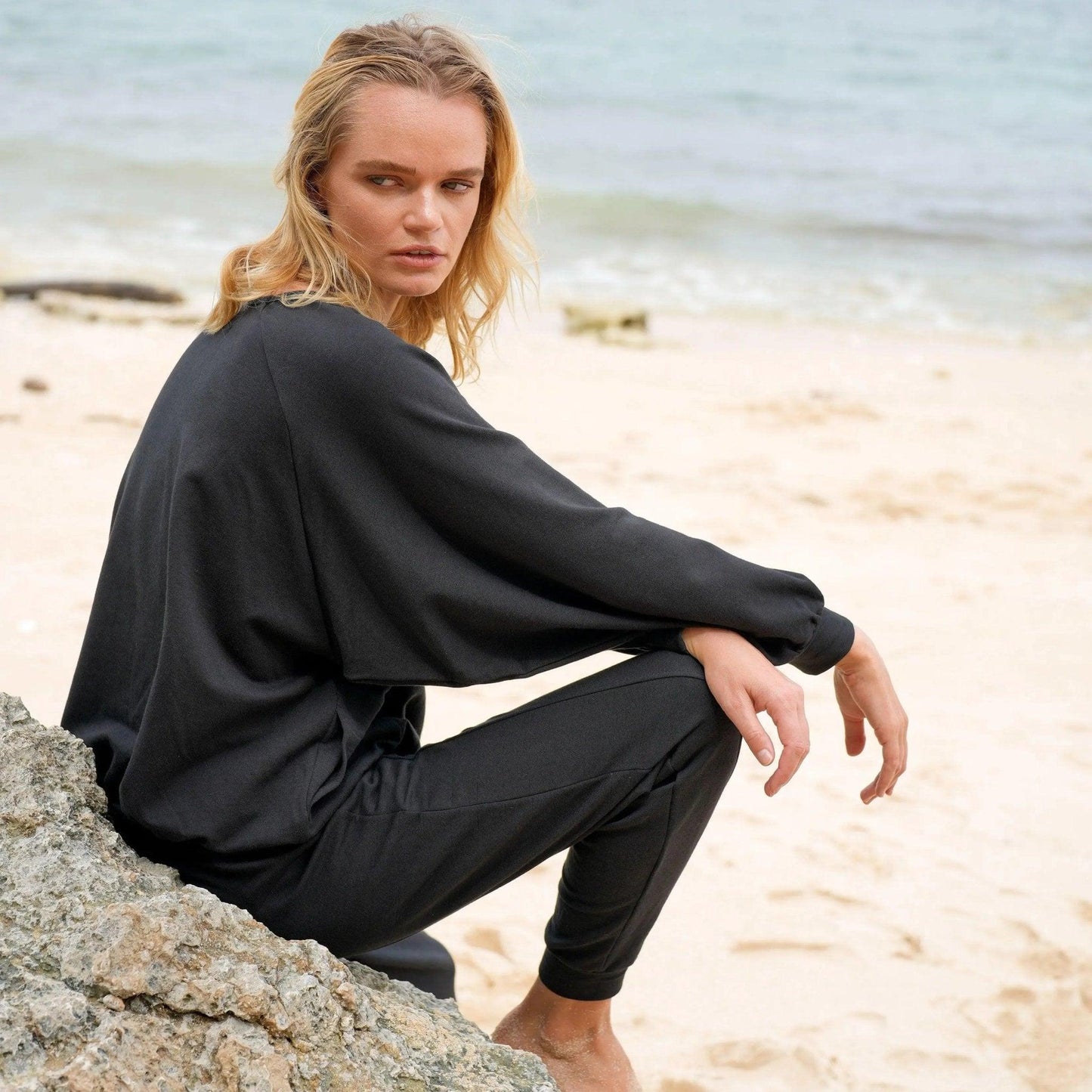 Woman in black long sleeve top and pants sitting on beach rock, coastal fashion outfit