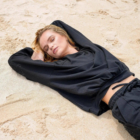 Woman in black hoodie and joggers relaxing on sandy beach, casual fashion outfit