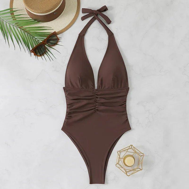 Brown halter neck one-piece swimsuit with ruched waist, straw hat, sunglasses, palm leaf on marble
