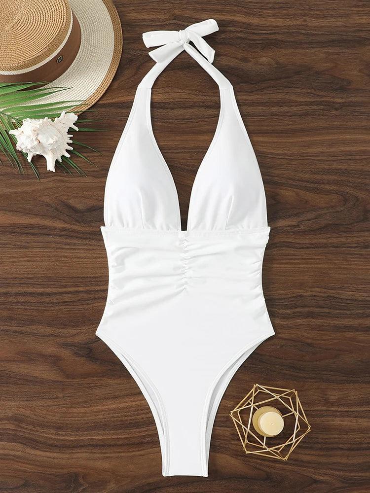 White halter one-piece swimsuit with ruched waist, beach hat, seashell, and gold decor on wood