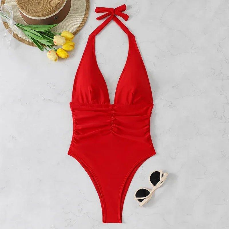 Red halter neck one-piece swimsuit with ruched detail, straw hat, yellow tulips, and sunglasses on marble