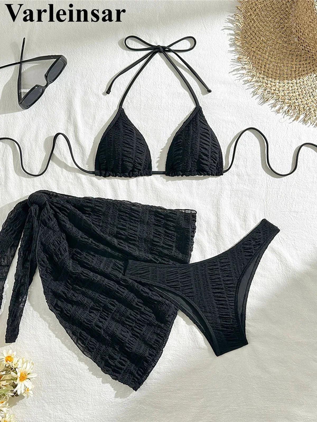 Black textured bikini set with tie-front top, matching bottoms, and sarong, styled with sunglasses and straw hat for beach fashion.