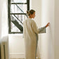 Woman in cozy linen robe with striped cuffs, standing by window in bright room