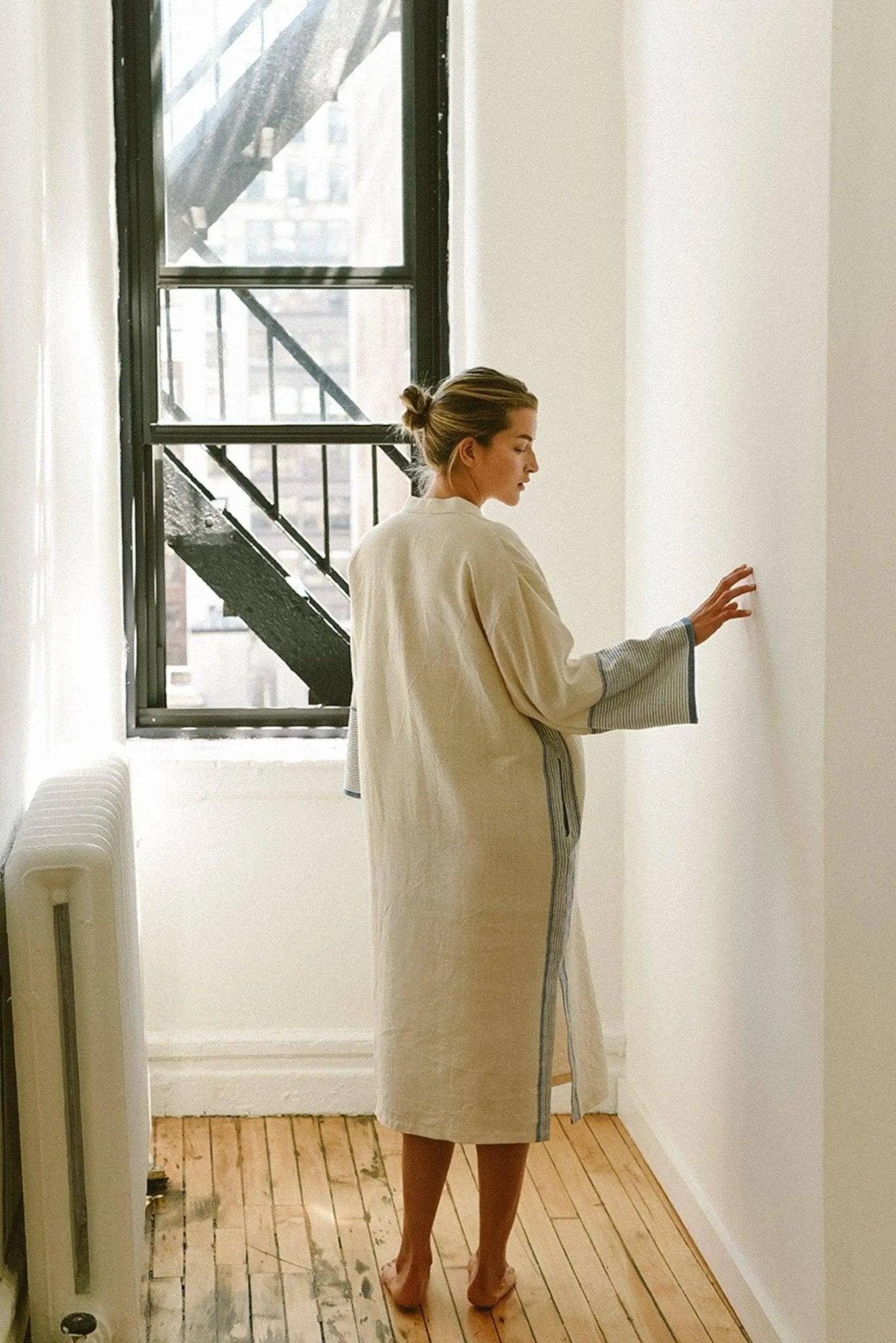 Woman in cozy linen robe with striped cuffs, standing by window in bright room