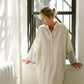 Woman in white oversized tunic dress with striped sleeves, standing by window in natural light