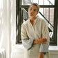 Woman in white linen robe with striped cuffs, standing by window, fashion lifestyle