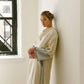 Woman in elegant cream robe with striped cuffs and side slit, leaning against white wall near window