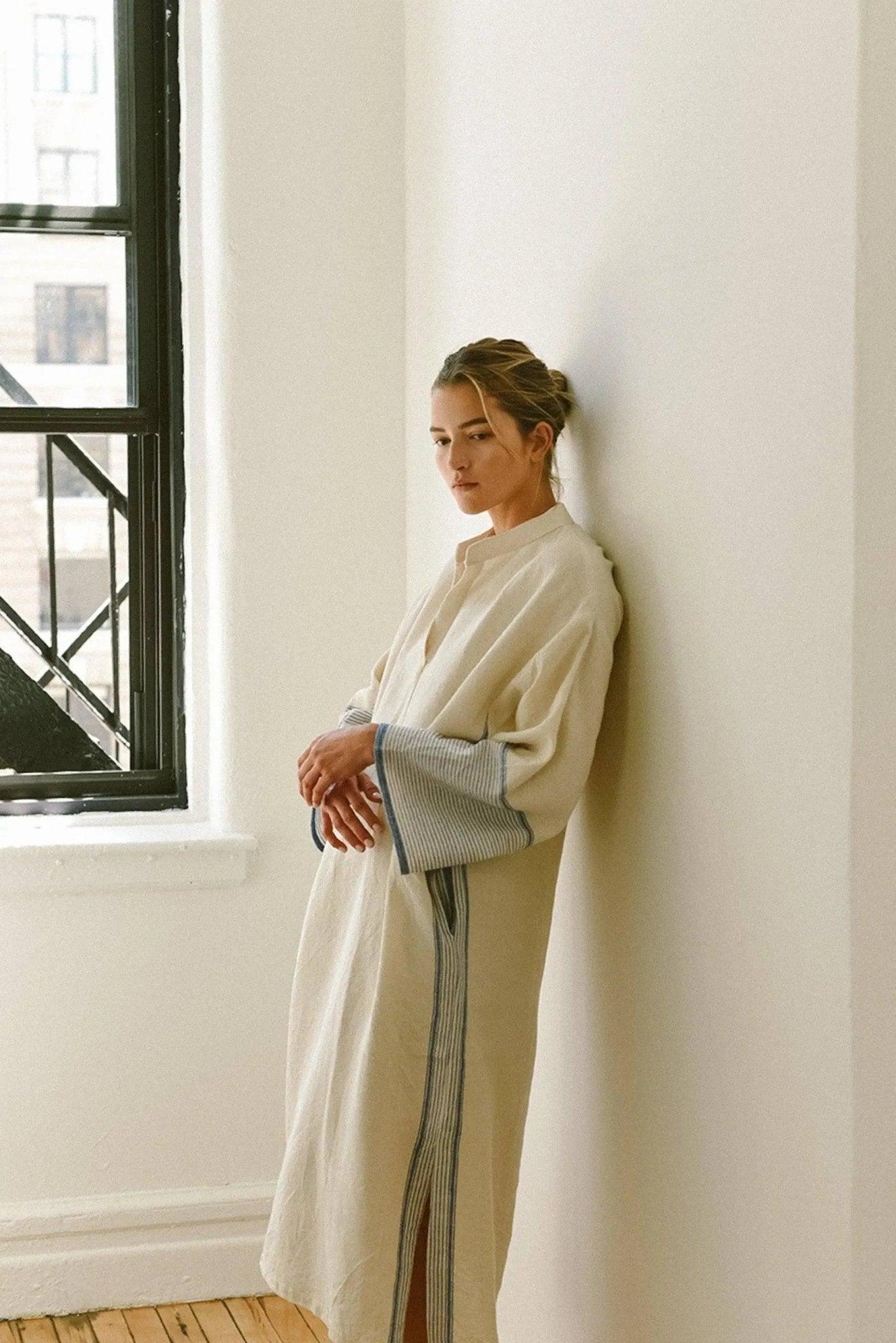 Woman in elegant cream robe with striped cuffs and side slit, leaning against white wall near window