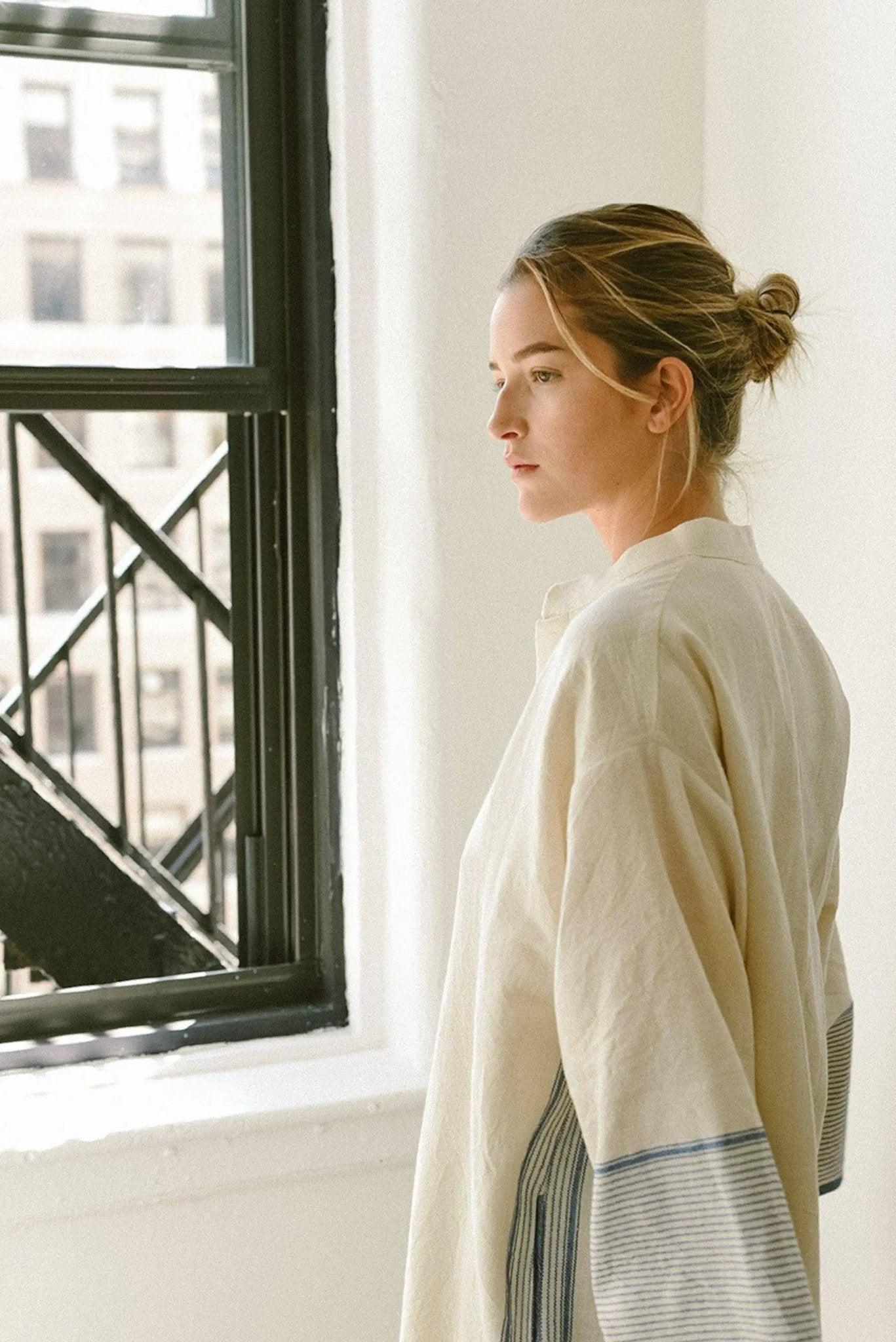 Woman in cream oversized shirt with striped sleeves, standing by window, minimalist fashion style
