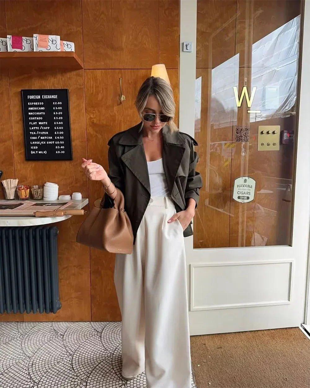 Woman in olive trench coat, white top, wide-leg pants, sunglasses, and tan handbag at café