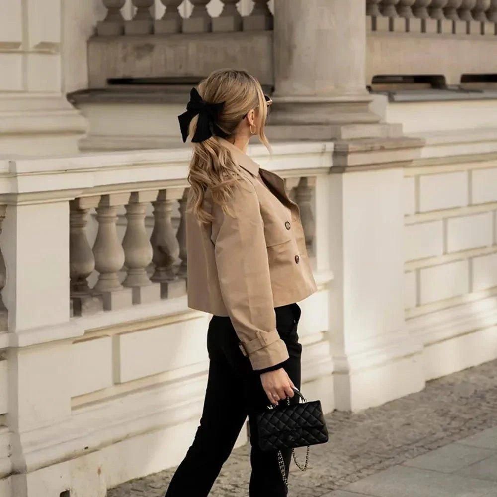 Woman in beige trench coat with black bow hair accessory, carrying quilted handbag, stylish street fashion