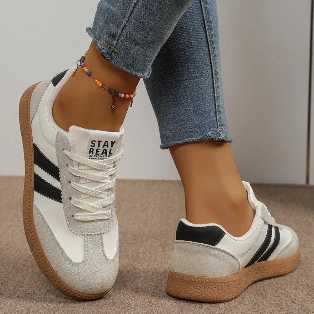 White and black women's sneakers with 'STAY REAL' text, paired with denim jeans and colorful anklet