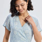 Woman in light blue floral wrap dress with V-neck and short sleeves, accessorized with gold necklace and earrings
