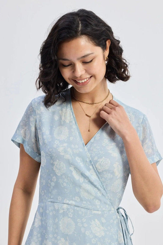 Woman in light blue floral wrap dress with V-neck and short sleeves, accessorized with gold necklace and earrings