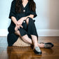 Woman in black dress with patterned slip-on shoes, sitting on woven pouf, fashion footwear