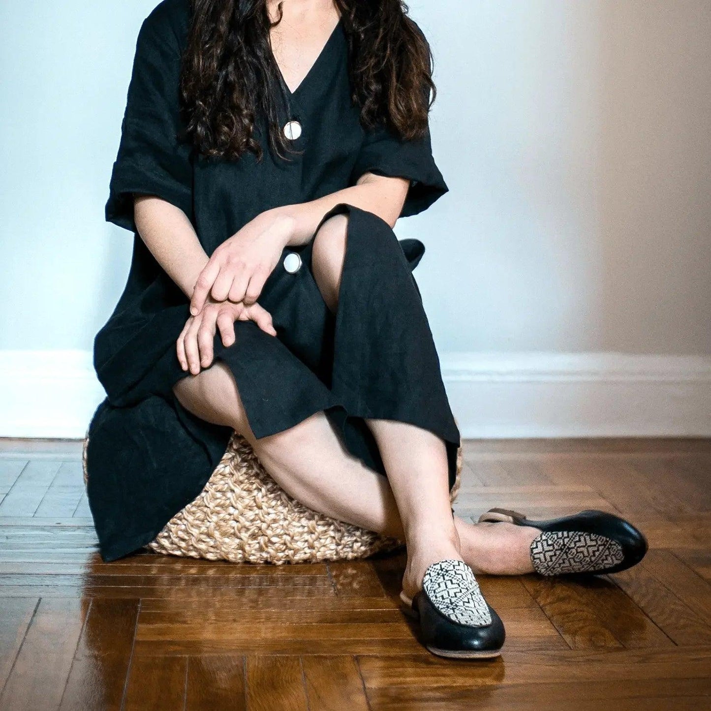 Woman in black dress with patterned slip-on shoes, sitting on woven pouf, fashion footwear