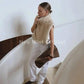 Woman in beige faux fur vest, white pants, holding clutch on modern staircase