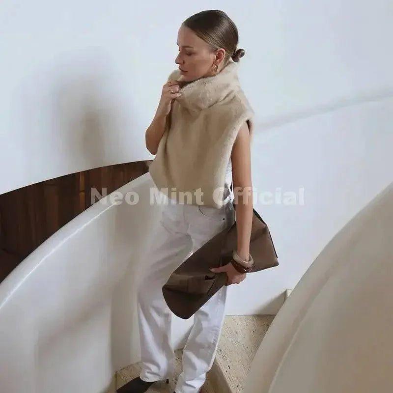 Woman in beige faux fur vest, white pants, holding clutch on modern staircase