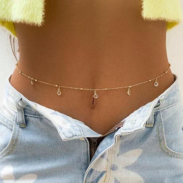 Gold chain belly button ring with dangling charms, worn with denim shorts and fuzzy top
