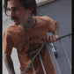 Man with tattoos wearing orange 'Raise Hell' graphic tee, leaning on metal railing