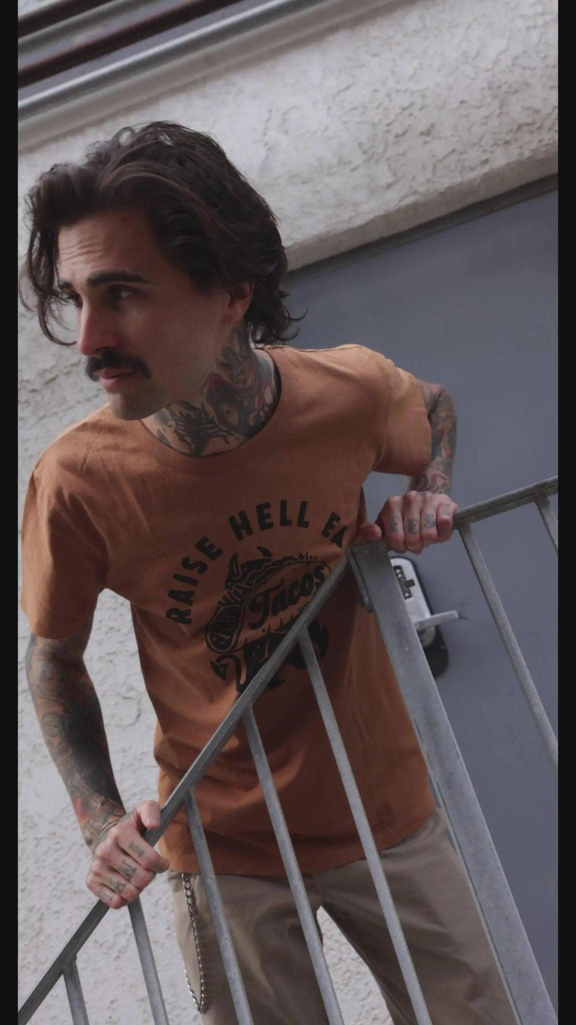 Man with tattoos wearing orange 'Raise Hell' graphic tee, leaning on metal railing