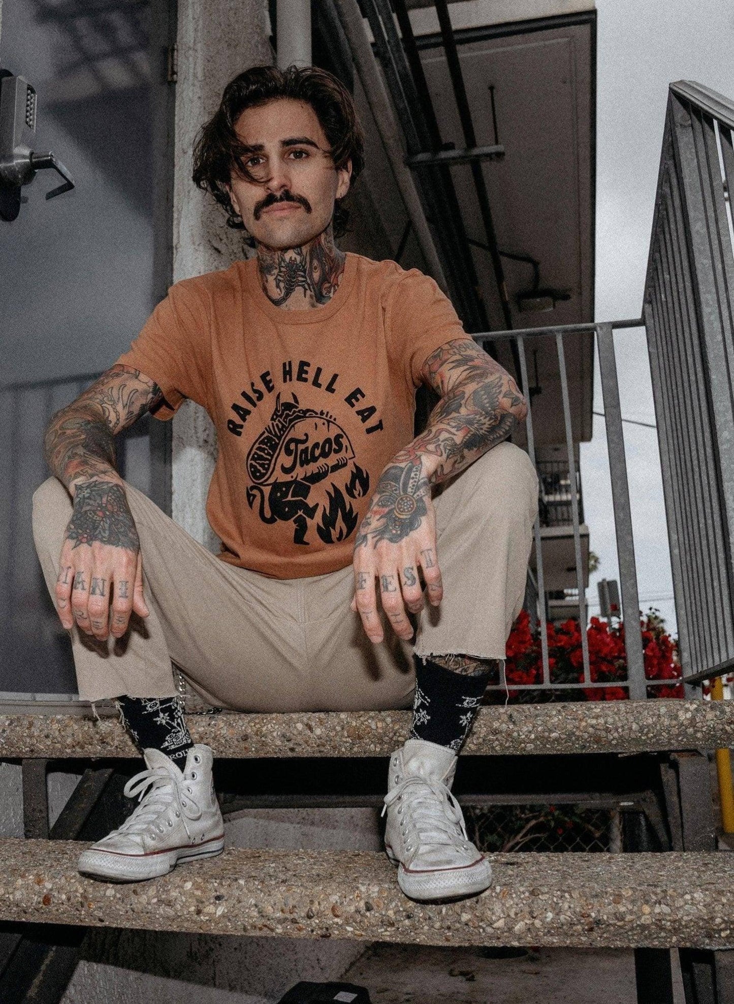 Man with tattoos wearing 'Raise Hell Eat Tacos' graphic tee, beige pants, and white sneakers on stairs