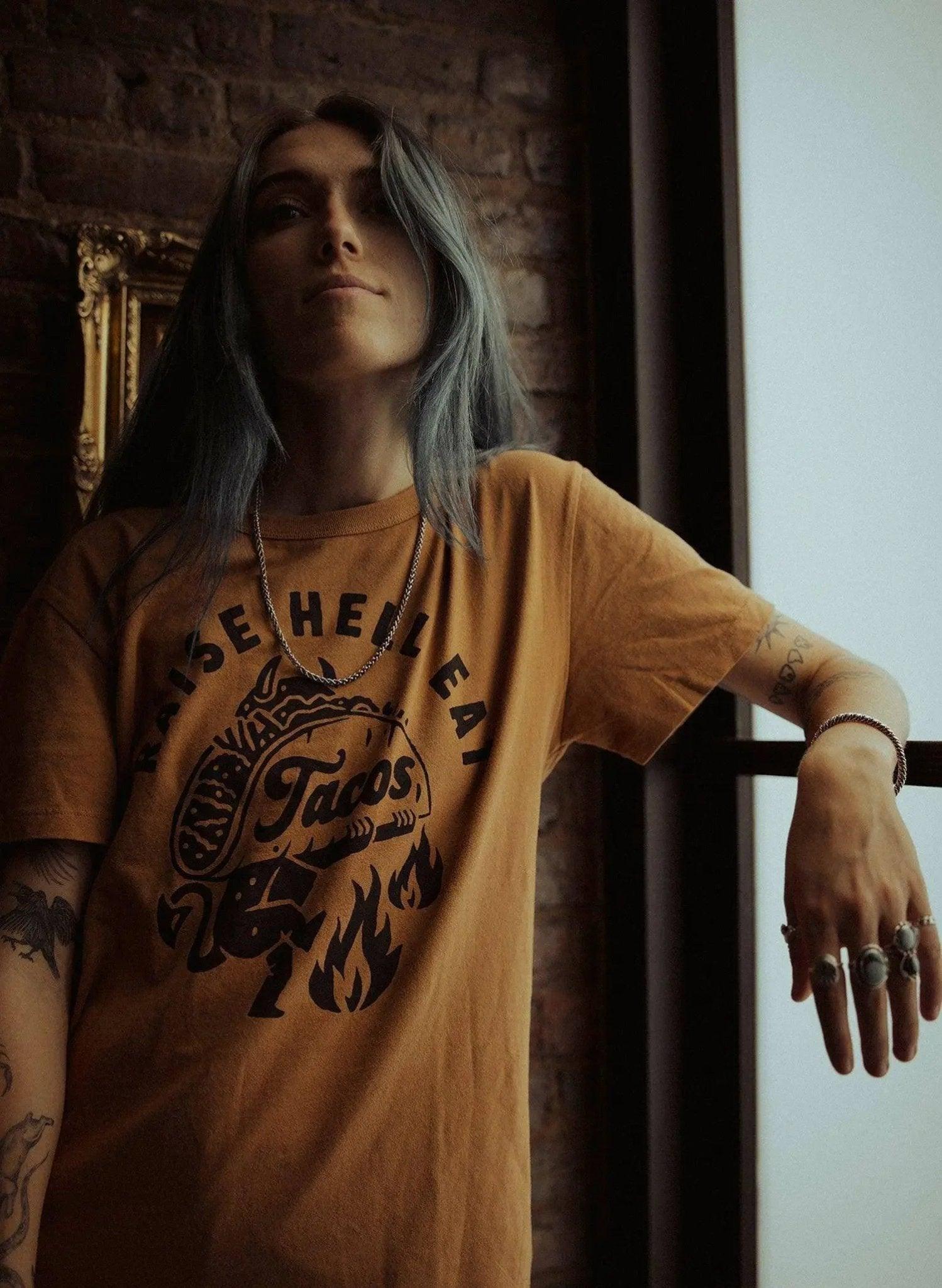 Woman wearing mustard graphic tee with 'Raise Hell Eat Tacos' print, tattoos, rings, and chain necklace