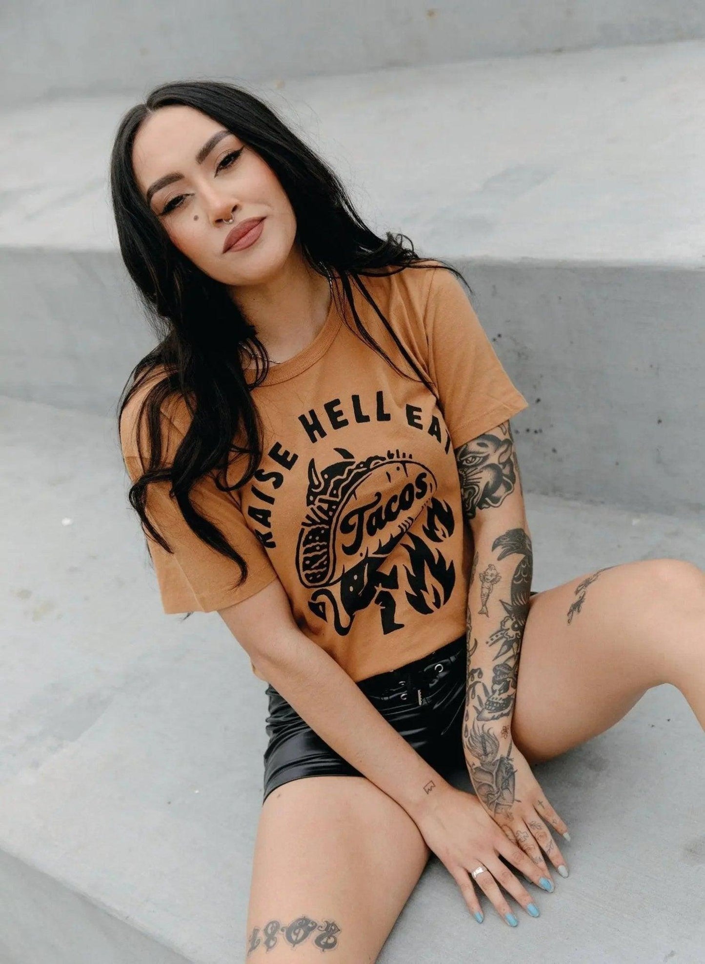 Woman in 'Raise Hell Eat Tacos' graphic tee, tattooed arms, black shorts, urban fashion style