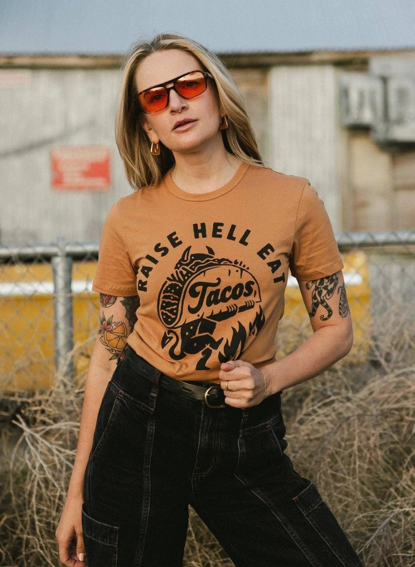 Woman wearing 'Raise Hell Eat Tacos' graphic tee, sunglasses, and black jeans, edgy street style fashion