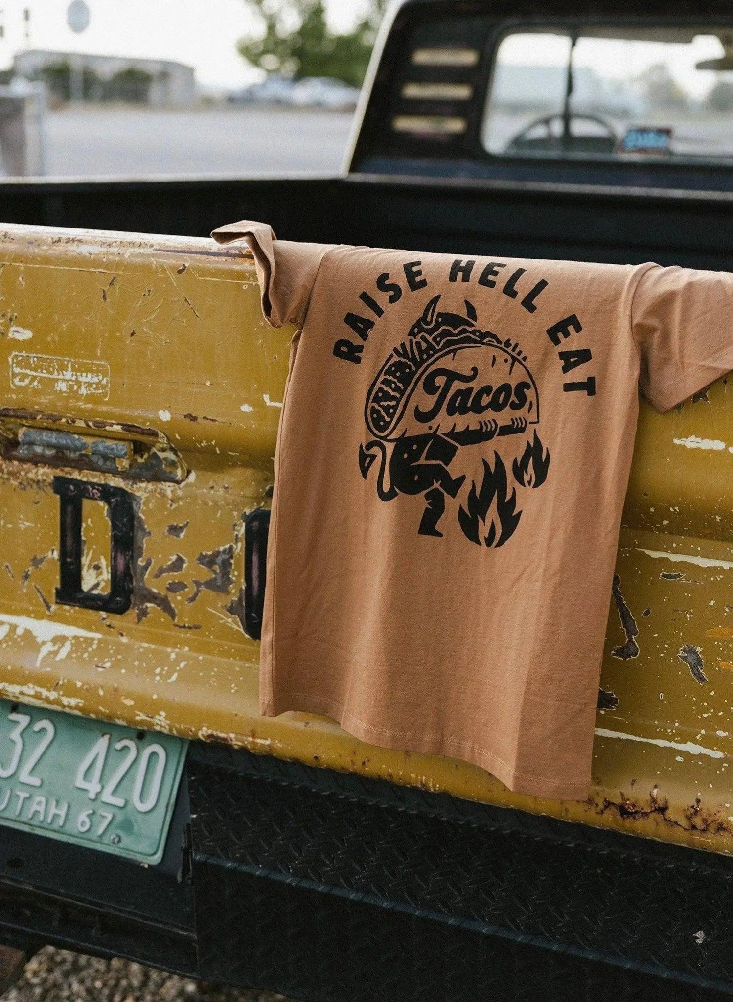 Tan graphic tee with 'Raise Hell Eat Tacos' design draped on vintage yellow truck tailgate