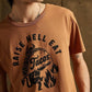 Brown graphic tee with 'Raise Hell Eat Tacos' design, featuring a taco with flames, worn by model.
