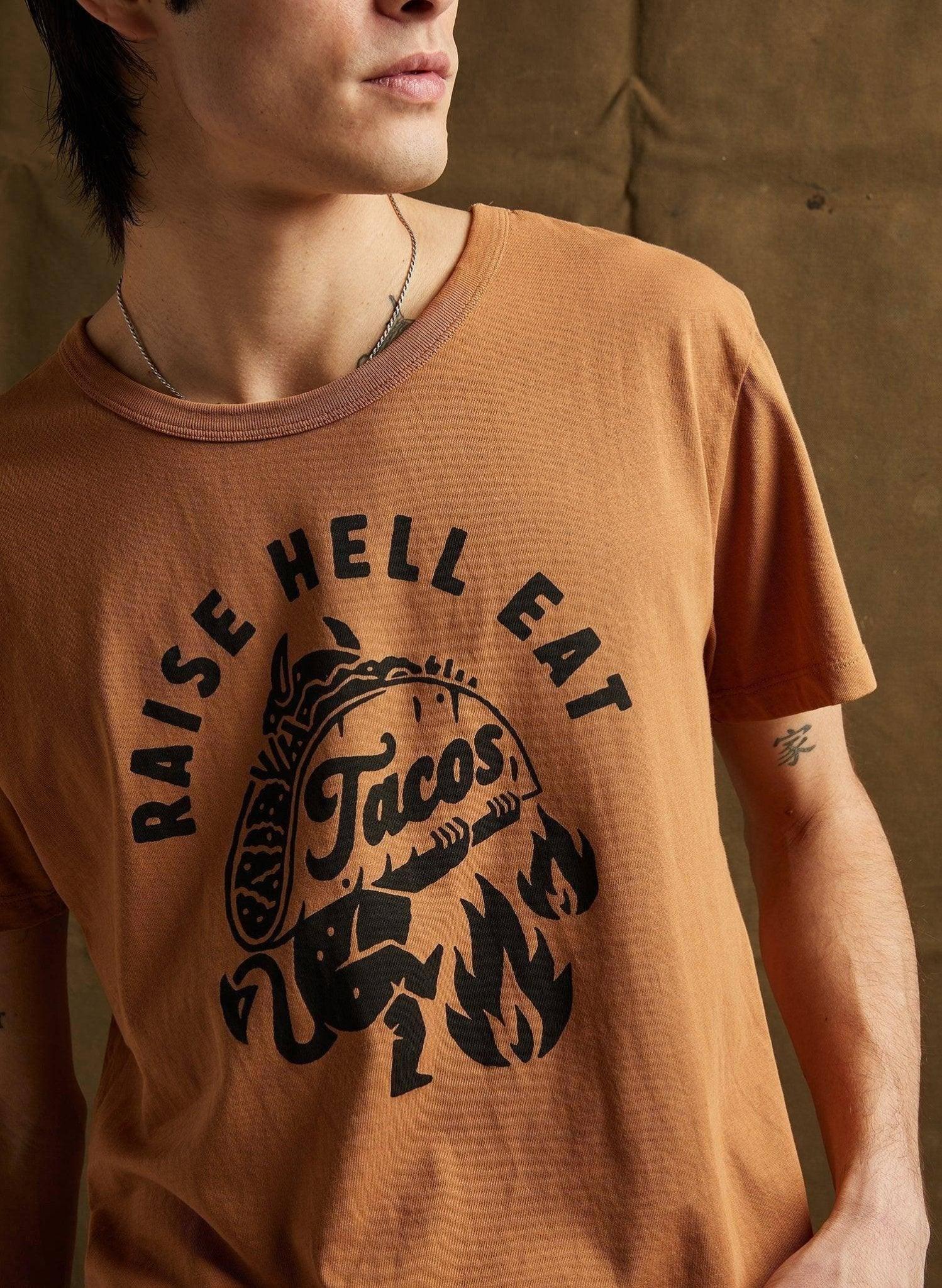 Brown graphic tee with 'Raise Hell Eat Tacos' design, featuring a taco with flames, worn by model.