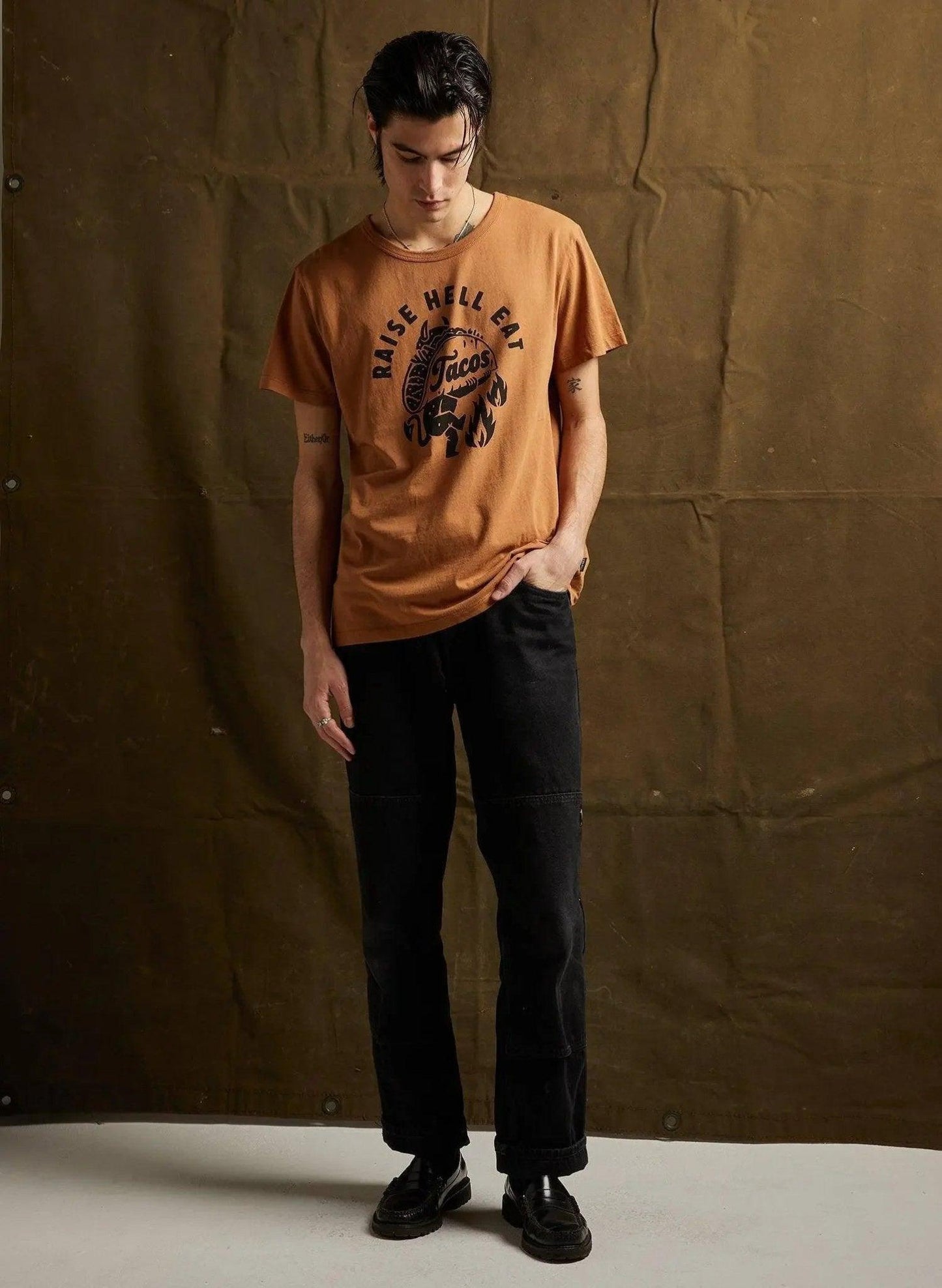 Man wearing orange graphic tee with 'Raise Hell Eat Tacos' print, black jeans, and loafers