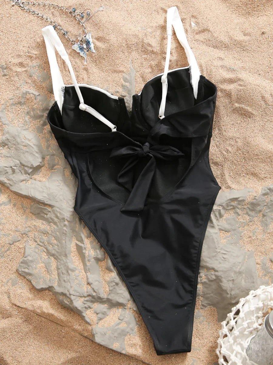 Black one-piece swimsuit with white straps and front bow, styled on sandy beach with butterfly necklace